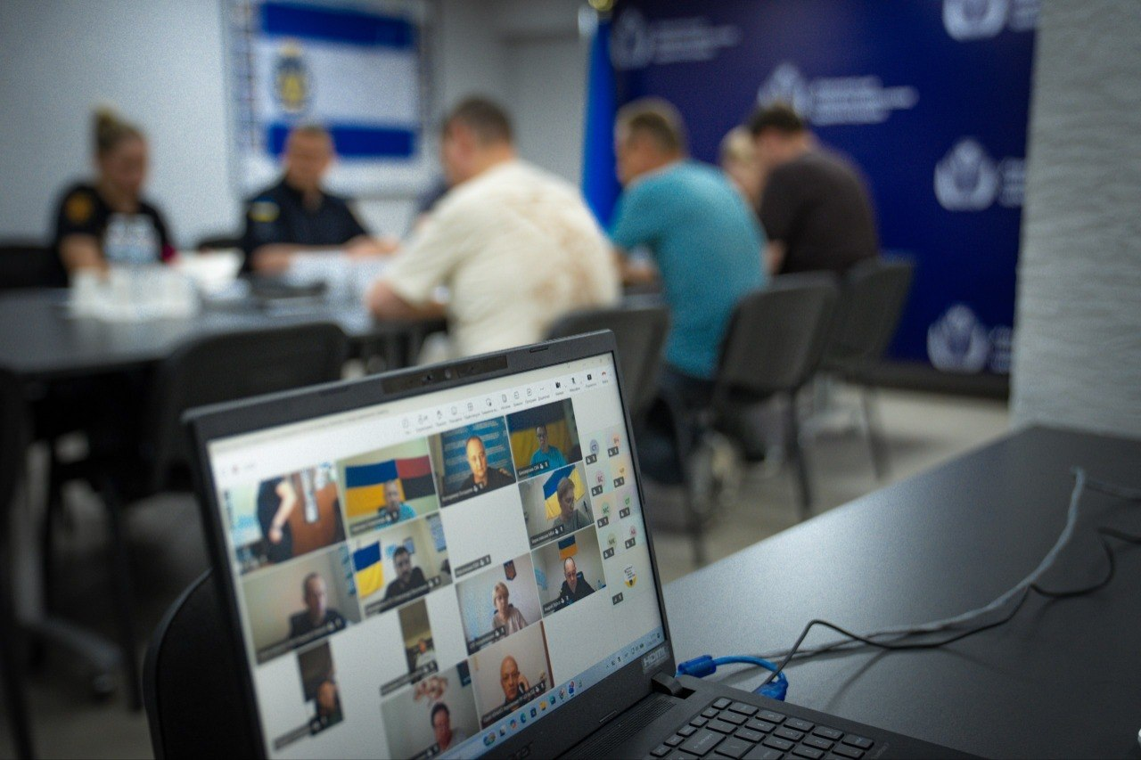 На передньому плані видно екран ноутбука із вікнами учасників онлайн-підключення під час наради щодо готовності укриттів. На задньому плані в розмитті (боке) сидять присутні в залі учасники зустрічі за довгим столом на тлі синього банера з логотипами ХОВА.