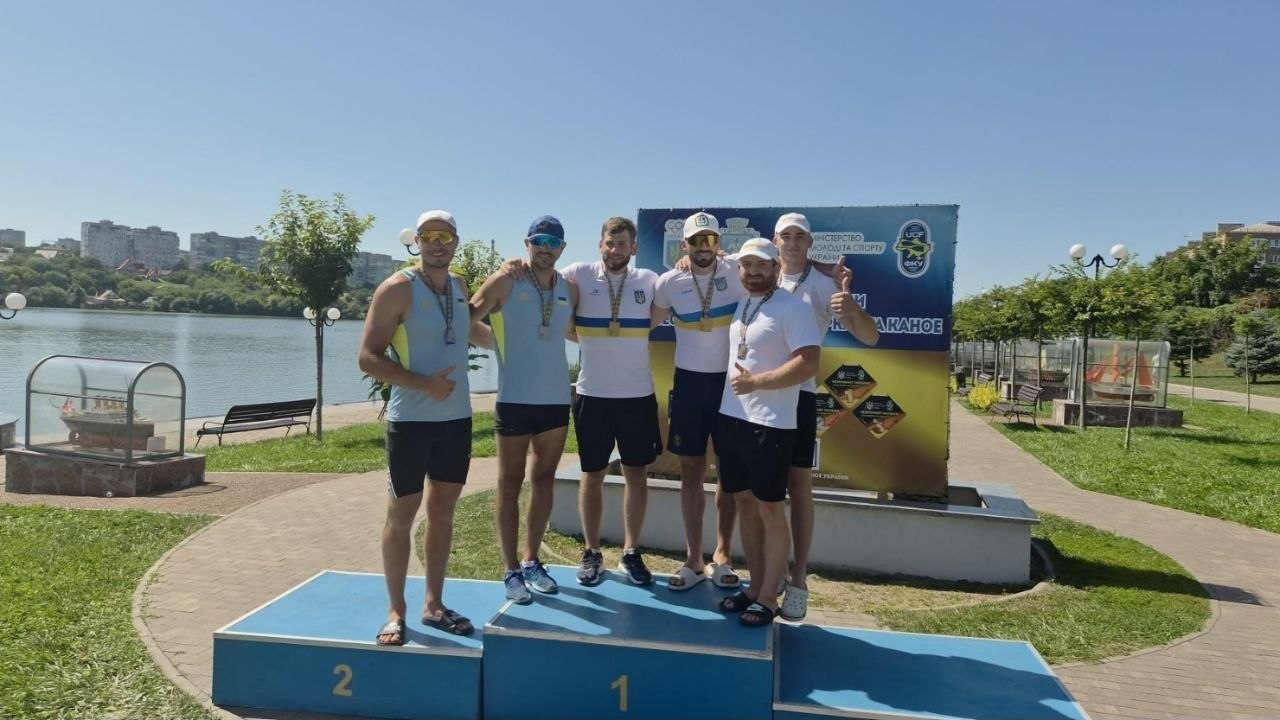 Нагородження призерів Чемпіонату України з веслування на байдарках і каное в Умані. Шість спортсменів-веслувальників із медалями на шиях стоять на п'єдесталі пошани (сходинки 1 та 2) на тлі офіційного банера змагань. На задньому плані видно водойму, набережну та міські будинки вдалині.