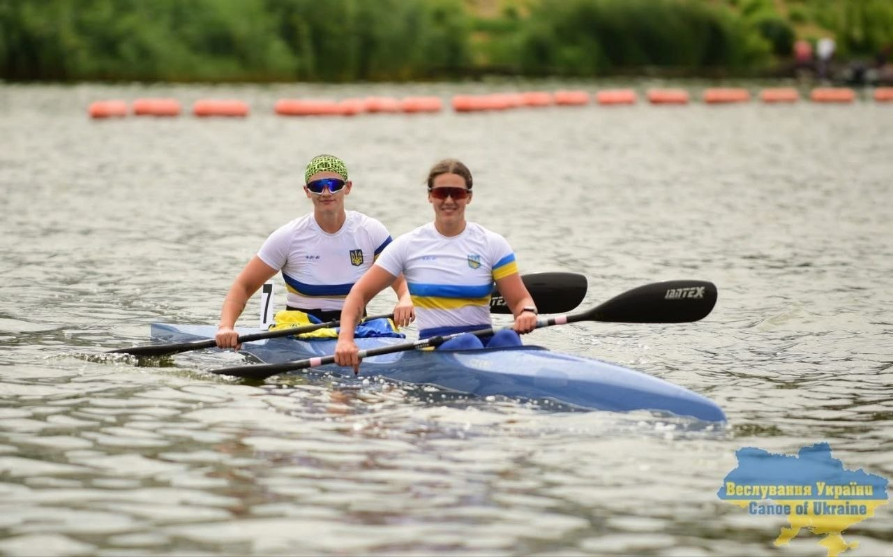 Дві спортсменки-веслувальниці у спортивній формі збірної України та сонцезахисних окулярах сидять у двомісній байдарці на воді, тримаючи в руках весла. На задньому плані видно помаранчеві обмежувальні буї на водній доріжці каналу, а у правому нижньому кутку — логотип ФВУ «Веслування України / Canoe of Ukraine».