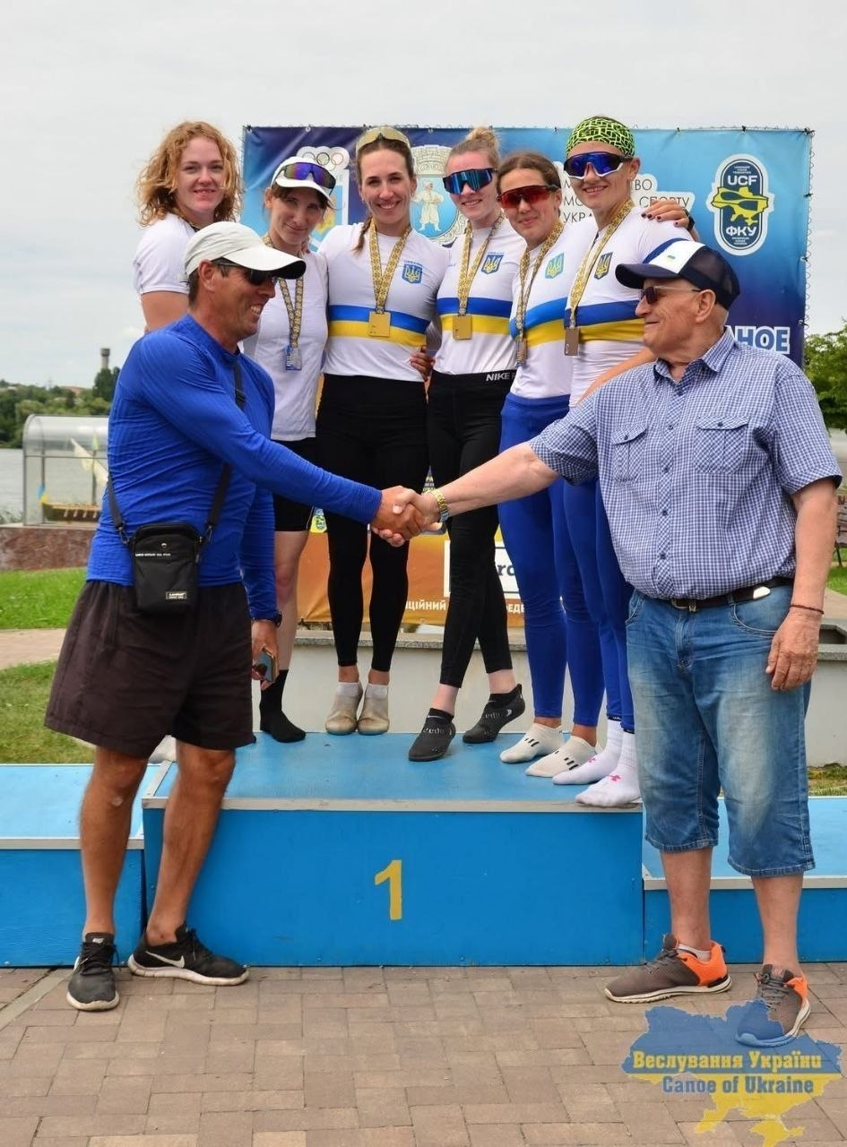 Церемонія нагородження на Чемпіонаті України з веслування. На першій сходинці п'єдесталу пошани стоять п'ять спортсменок у біло-синій формі збірної з медалями на шиях. На передньому плані перед п'єдесталом двоє чоловіків у головних уборах і сонцезахисних окулярах тиснуть один одному руки. На задньому плані — офіційний банер змагань.