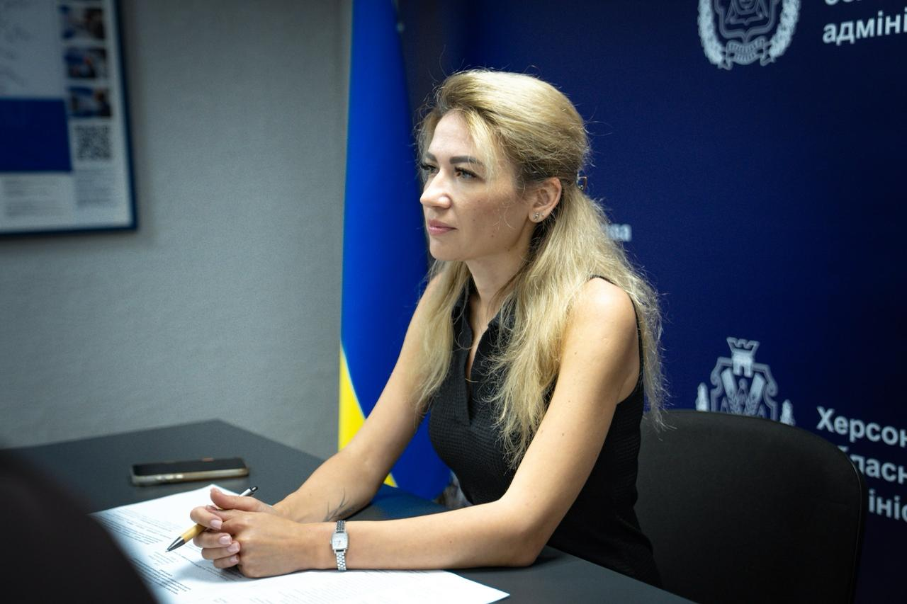 Заступник начальника Херсонської ОВА Ольга Малярчук головуюча на робочій нараді з питань пожежної безпеки населення. Світловолоса посадовиця в чорній блузці сидить за робочим столом із документами та ручкою на тлі прапора України та синьої стіни з символікою Херсонської обласної державної адміністрації.