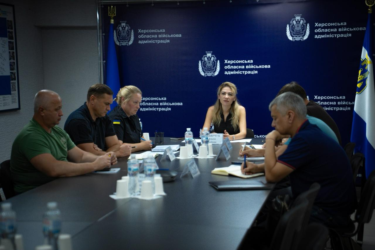 Заступник начальника Херсонської ОВА Ольга Малярчук проводить робочу нараду з питань пожежної безпеки населення. За великим столом із документами сидять заступник начальника Херсонської РВА Михайло Жадан (у зеленій футболці), представники ДСНС та ХОДА на тлі синього брендованого банера Херсонської обласної військової адміністрації.