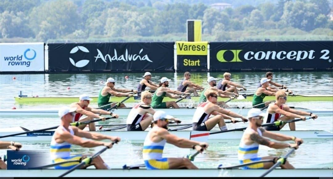 Екіпажі веслувальників на лінії старту «Vareze Start» під час Чемпіонату Європи з академічного веслування в Італії. На передньому плані — українські спортсмени у синьо-жовтій формі на човні з маркуванням екіпажу. На паралельних доріжках позаду готуються до старту суперники в зеленій та чорно-білій формі на тлі рекламних банерів «world rowing» та «concept 2».