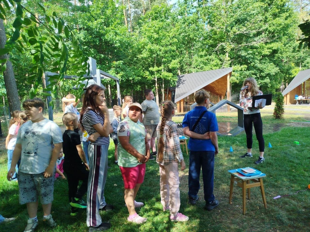 Заняття для дітей із безпеки в лісі від центру «Вільні разом» та ДП «Ліси України». На галявині біля дерев'яних альтанок ведуча з мікрофоном та папкою в руках звертається до групи вихованців табору «Перлина». Діти різного віку стоять на зеленому газоні під деревами та уважно слухають завдання інтерактивного заходу.