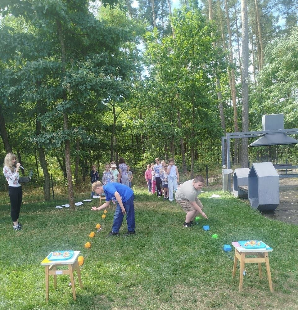 Діти беруть участь у спортивній естафеті під час заходу «Вікно в ліс: безпека дітей» у Житомирі. На лісовій галявині двоє хлопчиків наввипередки виконують завдання на газоні, збираючи кольорові предмети біля стільчиків. Інші вихованці табору «Перлина» стоять у черзі та вболівають, а ведуча ліворуч коментує гру в мікрофон.