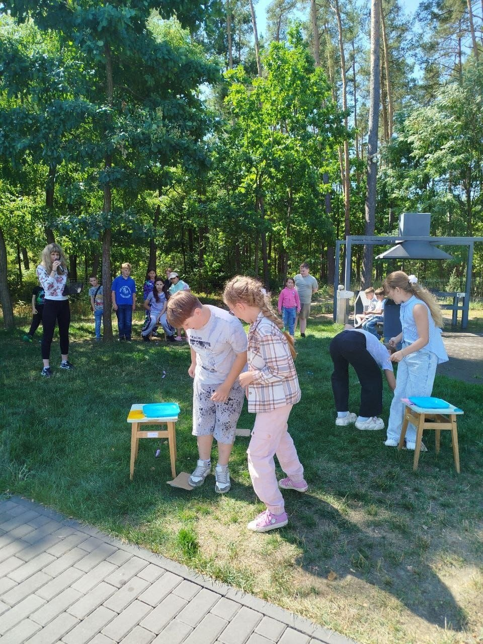 Командна естафета для дітей у межах безпекового проєкту «Вікно в ліс» у Житомирі. На парковій галявині біля дерев хлопчик та дівчинка виконують ігрове завдання біля стільчиків із блакитними тарілками. Позаду інші вихованці табору спостерігають за грою, а ведуча ліворуч тримає мікрофон і коментує змагання.