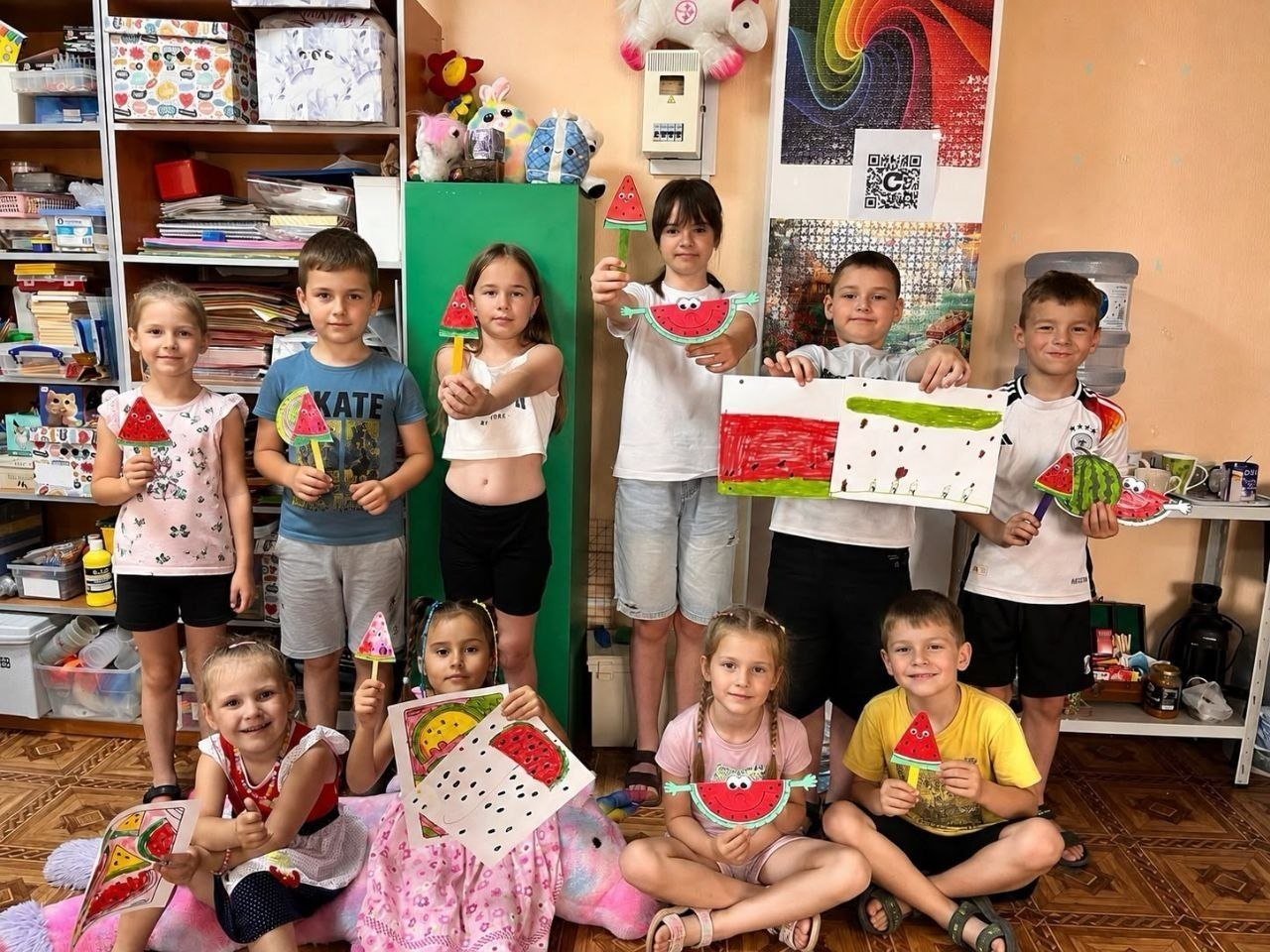 Групове фото дітей із родин ВПО з Херсонщини на арттерапевтичному занятті до Міжнародного дня кавуна в Миколаєві. Хлопчики та дівчата радісно позують у кімнаті, тримаючи в руках власноруч створені вироби: паперові аплікації, малюнки та саморобки у вигляді скибочок кавуна на паличках. Частина дітей стоїть, інші сидять на підлозі поруч із великою м'якою іграшкою.