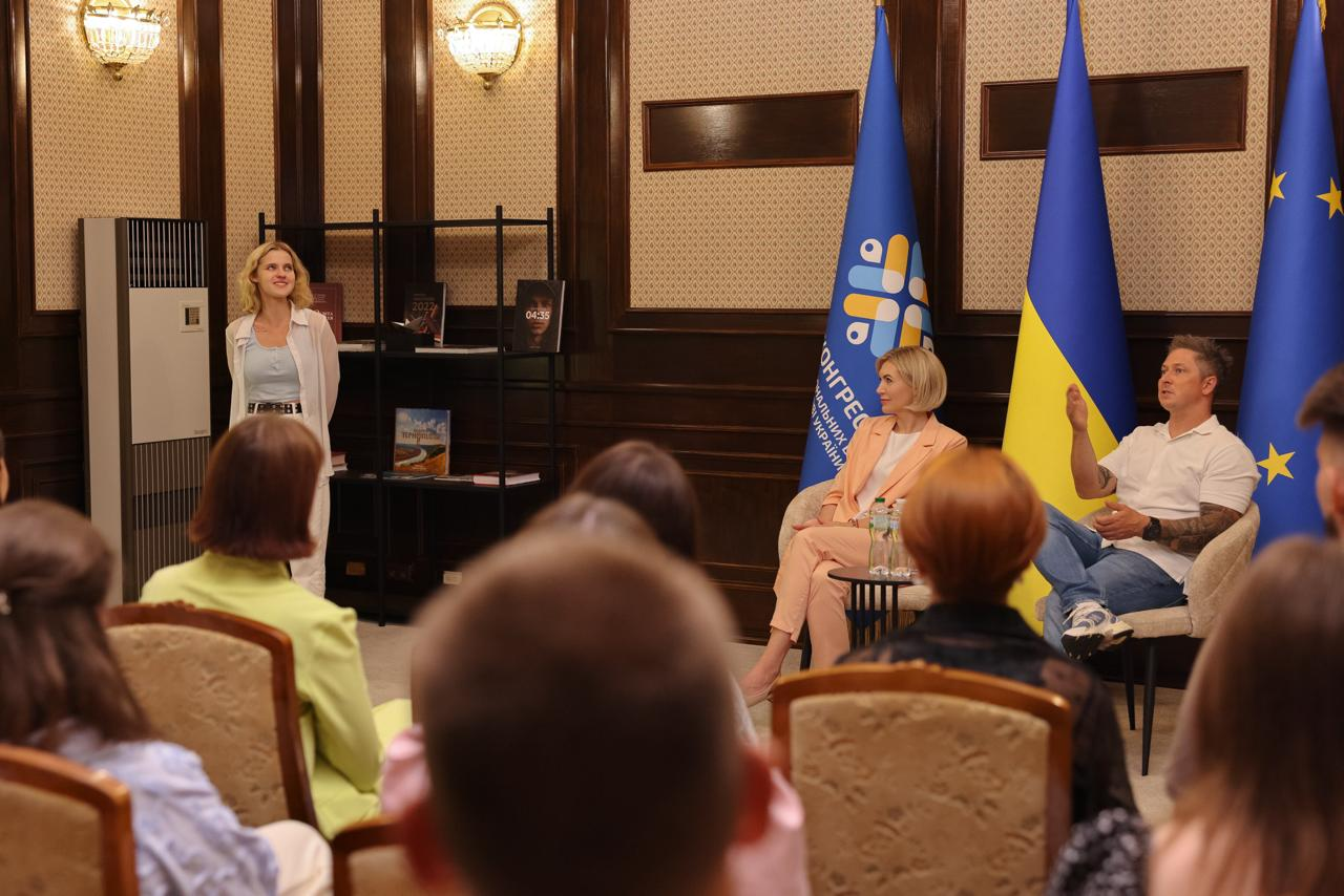 Засідання Палати регіональних молодіжних конгресів. Праворуч виступає чоловік у білій футболці, поруч із ним сидить жінка у персиковому костюмі на тлі прапорів України, ЄС та банера Конгресу. Ліворуч біля стелажа стоїть дівчина у світлому одязі. На передньому плані спиною до камери сидять учасники заходу, серед яких рудоволоса керівниця Молодіжного конгресу Херсонщини Катерина Саваєва.