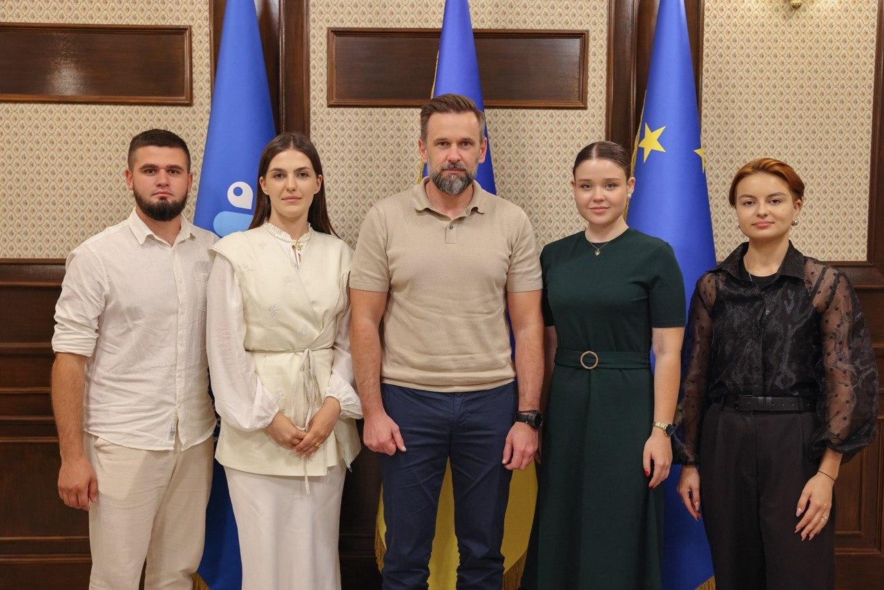 Групове фото учасників засідання Палати регіональних молодіжних конгресів. У центрі стоїть Заступник Керівника Офісу Президента України Віктор Микита у бежевій футболці-поло. Праворуч від нього — дівчина в темній сукні та керівниця Молодіжного конгресу Херсонщини Катерина Саваєва у чорній напівпрозорій блузці. Ліворуч — дівчина у світлому вбранні та молодий чоловік у білій сорочці. Позаду них розміщено державні прапори та банер Конгресу.