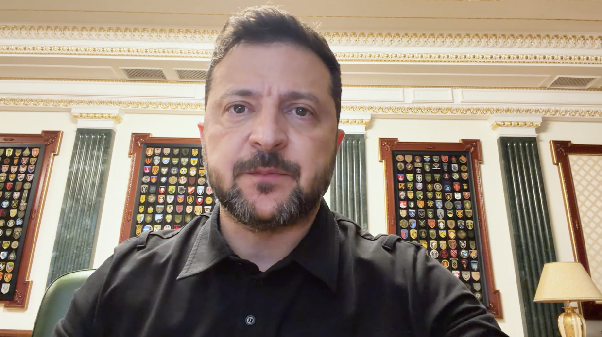 Президент України Володимир Зеленський у чорній сорочці виступає з відеозверненням на тлі офіційного кабінету зі стендами військових шевронів. Тема виступу присвячена важливості ППО для захисту життів та комунікації з міжнародними партнерами.