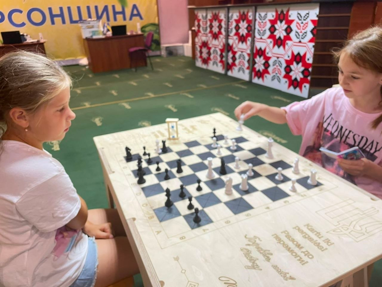 Дві дівчинки грають у шахи за світлим дерев'яним столом у координаційному центрі в Ужгороді. Одна дівчинка у рожевій футболці з принтом робить хід білою фігурою, а інша, у світло-рожевій футболці, зосереджено спостерігає за грою. На передньому плані столу вигравіювано герб України та напис «Разом до перемоги!», а поруч із шахівницею стоїть маленький пісочний годинник. На задньому плані приміщення на зеленій ковроліновій підлозі видно банер із написом «Херсонщина» та декоративні ширми з традиційним українським червоно-чорним орнаментом.