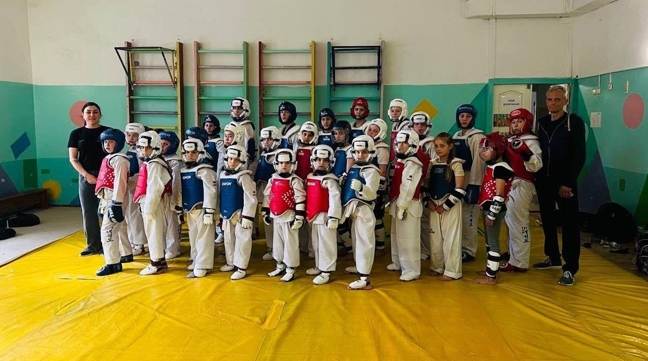 Велика група дітей-переселенців з Херсонщини у повному захисному спорядженні для тхеквондо (білі добки, різнокольорові жилети та шоломи) стоїть на жовтому маті для спільного фото. Зліва від дітей стоїть тренерка Мадонна Баранова, а справа — її колега-чоловік. На задньому плані світлої спортивної зали координаційного центру в Миколаєві видно різнокольорові шведські стінки та відчинені двері.