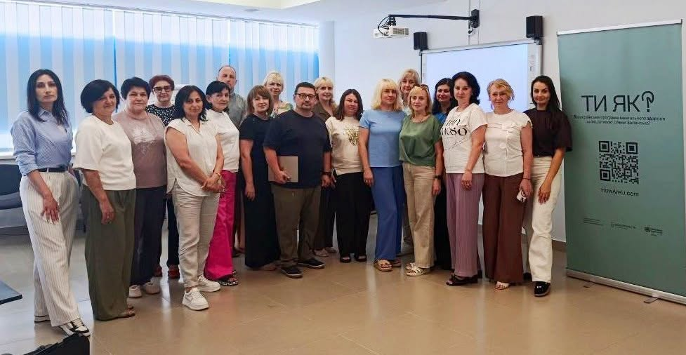 рупа працівників Центрів зайнятості стоїть у ряд для спільного фото у світлій залі під час тренінгу проєкту «Національна платформа самоосвіти». Справа від учасників розміщено вертикальний зелений банер Всеукраїнської програми ментального здоров'я «Ти як?» із QR-кодом. На задньому плані приміщення видно інтерактивну білу дошку з проєктором та велике вікно з жалюзі.