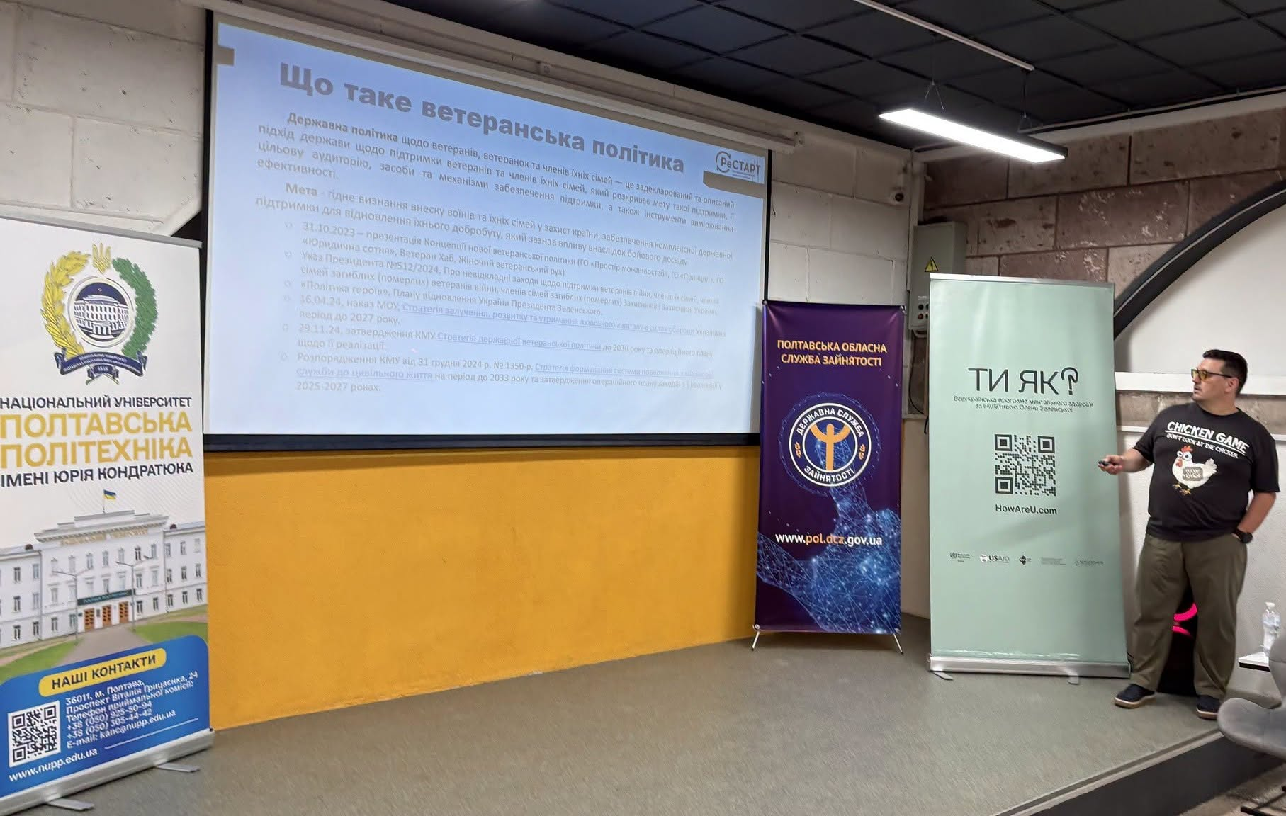 Чоловік у чорній футболці та окулярах виступає перед аудиторією в лекційній залі Полтавської політехніки, тримаючи в руці клікер. На великому настінному екрані за ним відображено слайд презентації з заголовком «Що таке ветеранська політика». Справа від екрана стоїть фіолетовий банер Полтавської обласної служби зайнятості та світло-зелений банер програми ментального здоров'я «Ти як?», а зліва — інформаційний стенд університету.