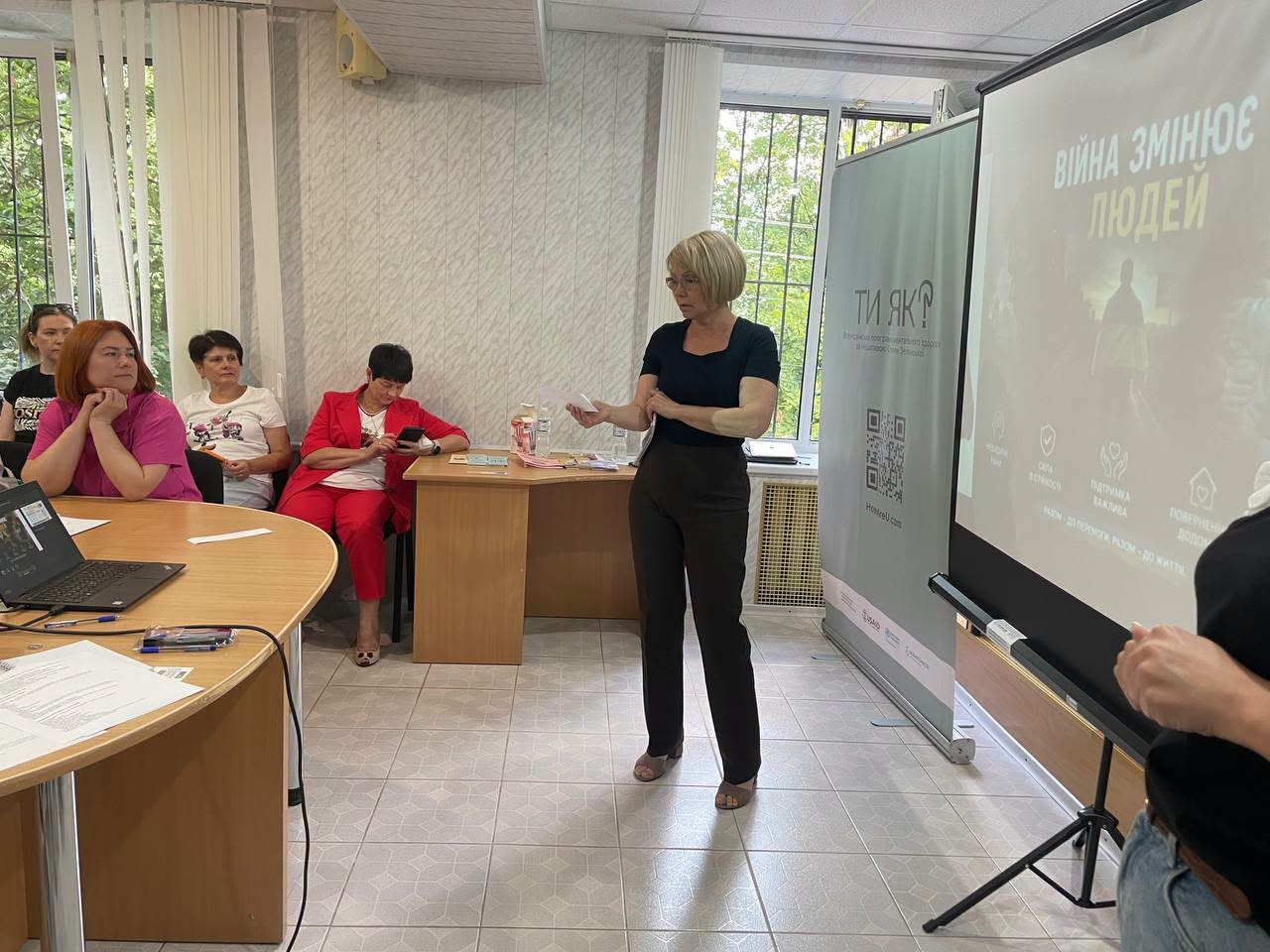 Жінка-спікерка з короткою світлою зачіскою виступає в залі перед аудиторією під час спеціалізованого тренінгу. Праворуч від неї на великому проєкційному екрані виведено слайд із написом «Війна змінює людей», а поруч стоїть банер програми ментального здоров'я «Ти як?». Ліворуч за столами сидять учасниці тренінгу, які слухають доповідачку.