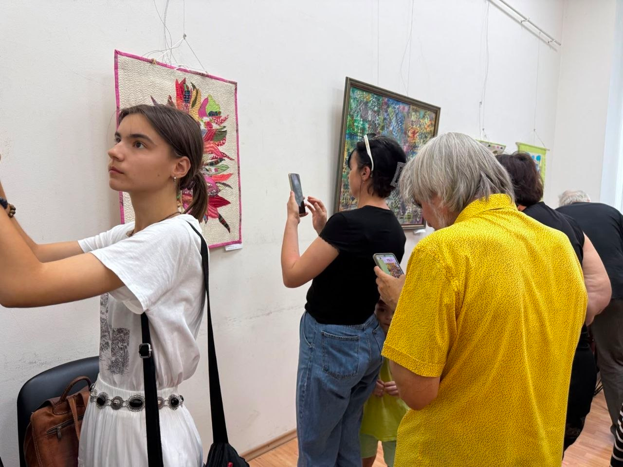 Відвідувачі виставки «Від витоків до сьогодення» в Одесі, серед яких переселенці з Херсонщини, розглядають експонати та фотографують їх на смартфони. На передньому плані ліворуч дівчина у білій сукні дивиться на виставлені роботи. На світлій стіні зали розвішані картини та текстильні панно, а чоловік у яскравій жовтій сорочці на передньому плані вивчає інформацію у своєму телефоні.