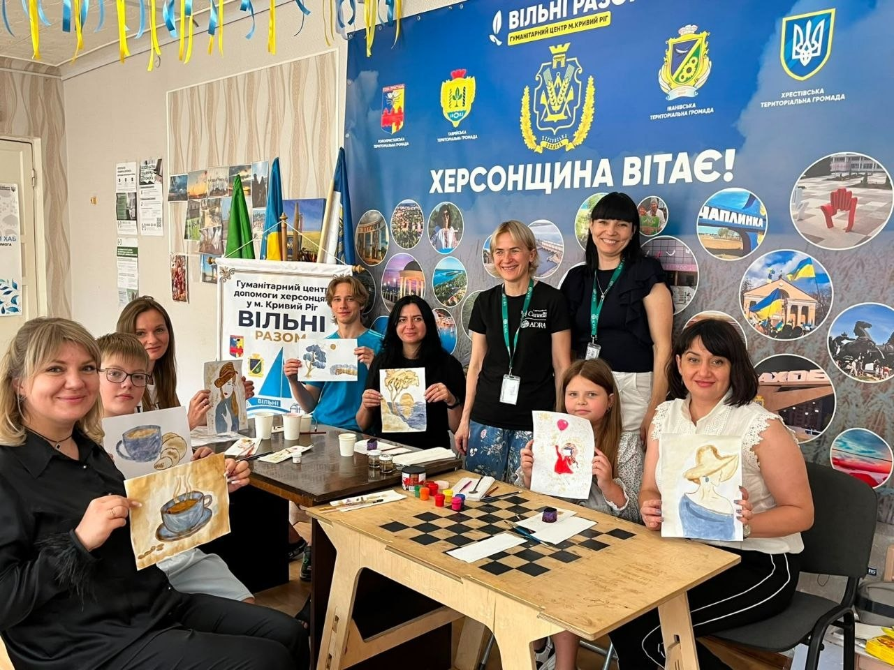 Група дітей та дорослих переселенців із Херсонщини сидить за столами у гуманітарному центрі «Вільні разом» у Кривому Розі. Учасники тримають у руках свої роботи, створені під час арттерапевтичного майстер-класу з малювання кавою «Малюємо ароматом». Позаду них стоять дві представниці БФ «АДРА» у чорних фірмових футболках. На задньому плані кімнати розміщено великий синій банер із написом «Херсонщина вітає!», гербами громад та фотоколажем.