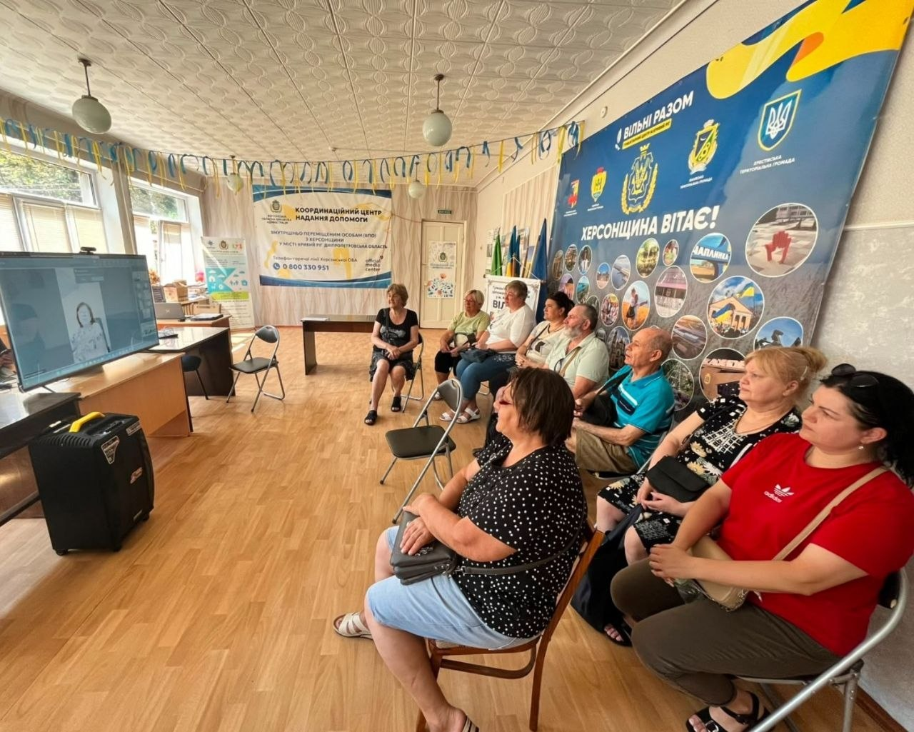 Група переселенців із Херсонщини сидить у залі координаційного центру «Вільні разом» у Кривому Розі під час онлайн-заходу, присвяченого історії місцевої геральдики. Люди зосереджено дивляться на великий монітор, де транслюється онлайн-зустріч. Праворуч на стіні розміщено великий синій банер із написом «Херсонщина вітає!», а на задньому плані видно світлий інформаційний стенд координаційного центру та державні прапори.