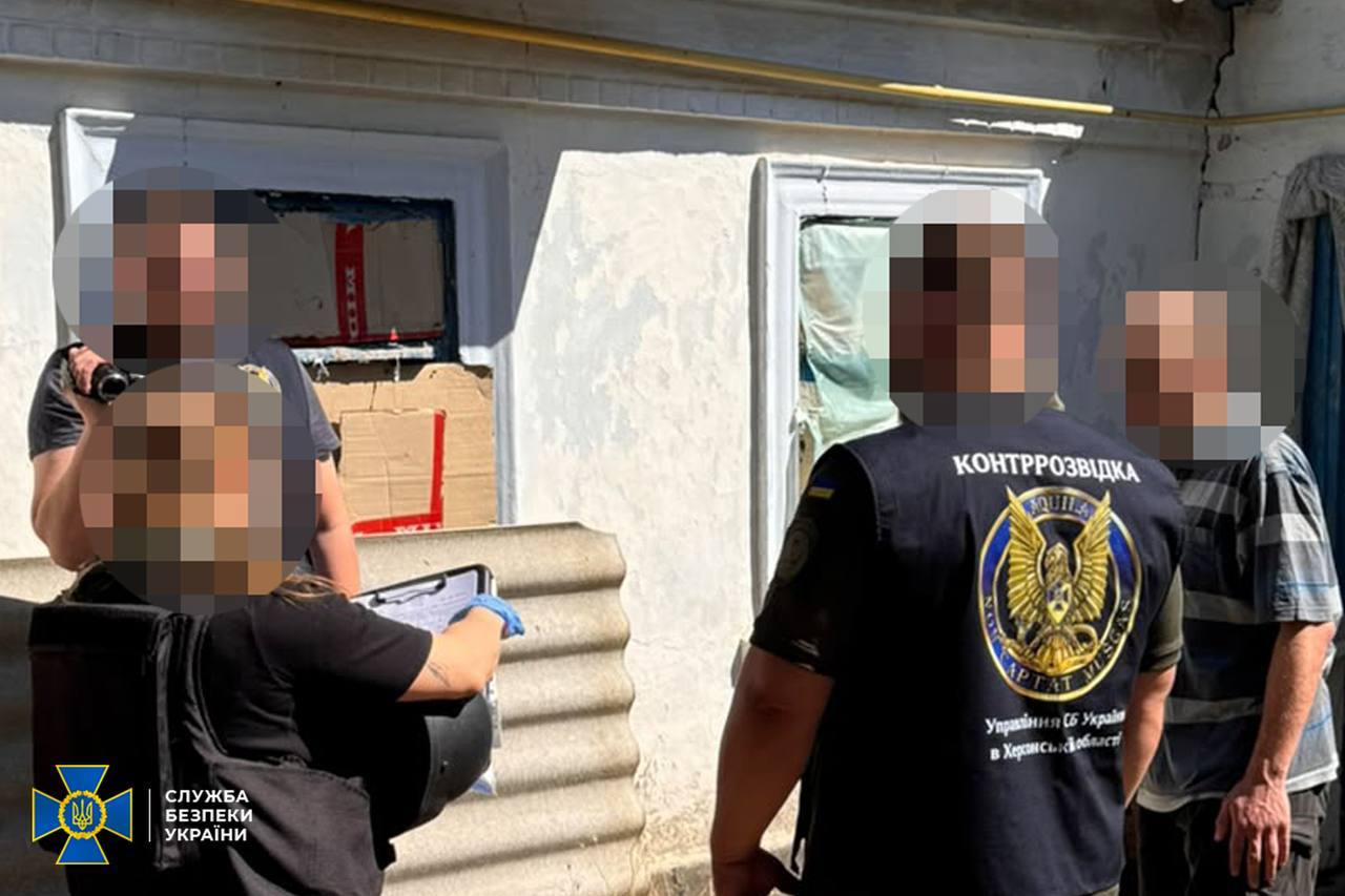 Співробітники контррозвідки СБУ в Херсонській області проводять слідчі дії біля житлового будинку в Херсоні в присутності підозрюваного чоловіка у смугастій футболці. На передньому плані праворуч спиною до камери стоїть правоохоронець у тактичному жилеті з написом «КОНТРОЗВІДКА» та великою круглою емблемою підрозділу УСБУ в Херсонській області. Інші співробітники ліворуч фіксують процес на відеокамеру та роблять записи у документах. Обличчя всіх присутніх повністю розмиті, а в лівому нижньому кутку розміщено офіційний логотип Служби безпеки України.