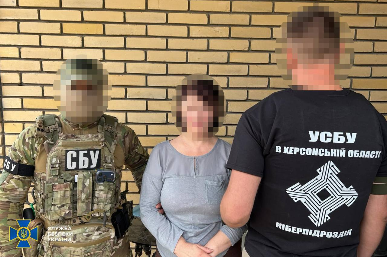 Співробітники СБУ затримують підозрювану в інтернет-агітації жінку у Херсоні на тлі жовтої цегляної стіни. Ліворуч стоїть оперативник у камуфляжній формі та плитоносці з написом «СБУ». Праворуч спиною до камери зафіксований співробітник у чорній футболці з написом «УСБУ В ХЕРСОНСЬКІЙ ОБЛАСТІ КІБЕРПІДРОЗДІЛ» та цифровою емблемою. Обличчя жінки та правоохоронця ліворуч повністю розмиті в інтересах слідства, а в нижньому кутку додано офіційний логотип Служби безпеки України.