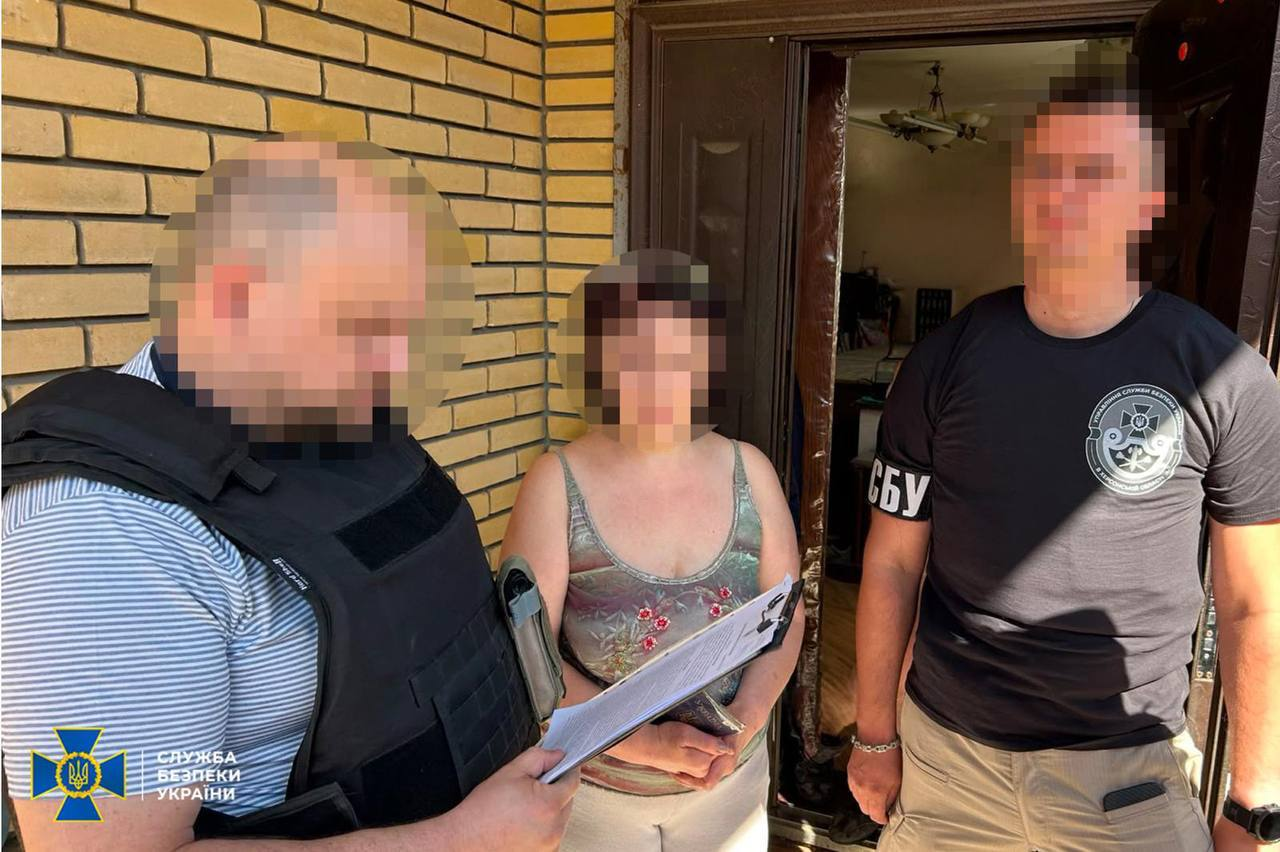 Двоє співробітників СБУ проводять слідчі дії з підозрюваною у прокремлівській агітації жінкою біля дверей будинку у Херсоні. На передньому плані ліворуч правоохоронець у чорному бронежилеті тримає папку з паперами та ознайомлює фігурантку з процесуальними документами. Праворуч стоїть оперативник у чорній футболці з білим написом «СБУ» та круглим шевроном відомства на тлі жовтої цегляної стіни. Обличчя жінки та обох правоохоронців повністю розмиті, а в лівому нижньому кутку додано офіційний логотип Служби безпеки України.