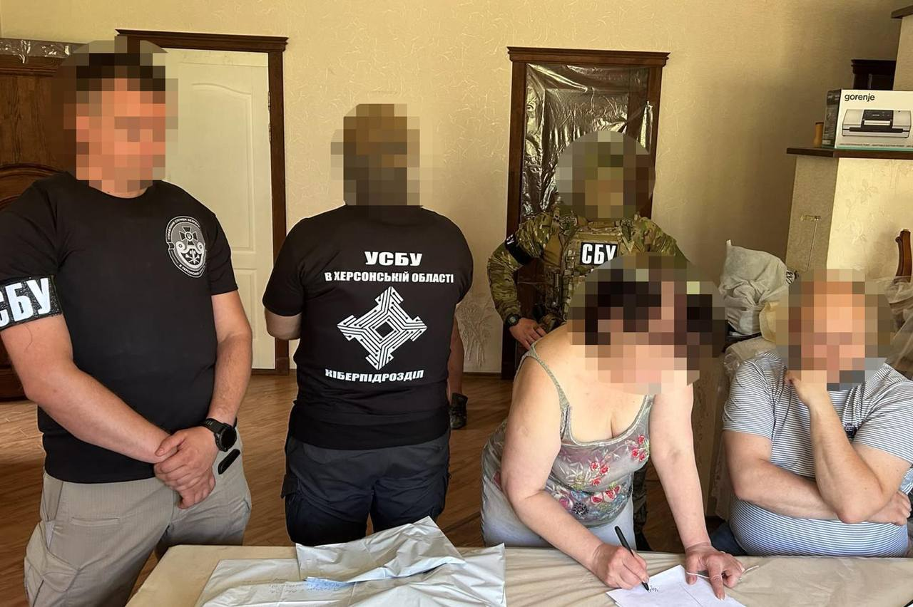 Троє співробітників СБУ проводять слідчі дії у кімнаті житлового будинку в Херсоні в присутності підозрюваних жінки та чоловіка. На передньому плані жінка підписує процесуальні документи за столом. Ліворуч стоїть оперативник у чорній футболці з логотипом відомства, по центру спиною до камери — співробітник у футболці з написом «УСБУ В ХЕРСОНСЬКІЙ ОБЛАСТІ КІБЕРПІДРОЗДІЛ», а на задньому плані — боєць у камуфляжі та бронежилеті «СБУ». Обличчя всіх присутніх повністю розмиті.