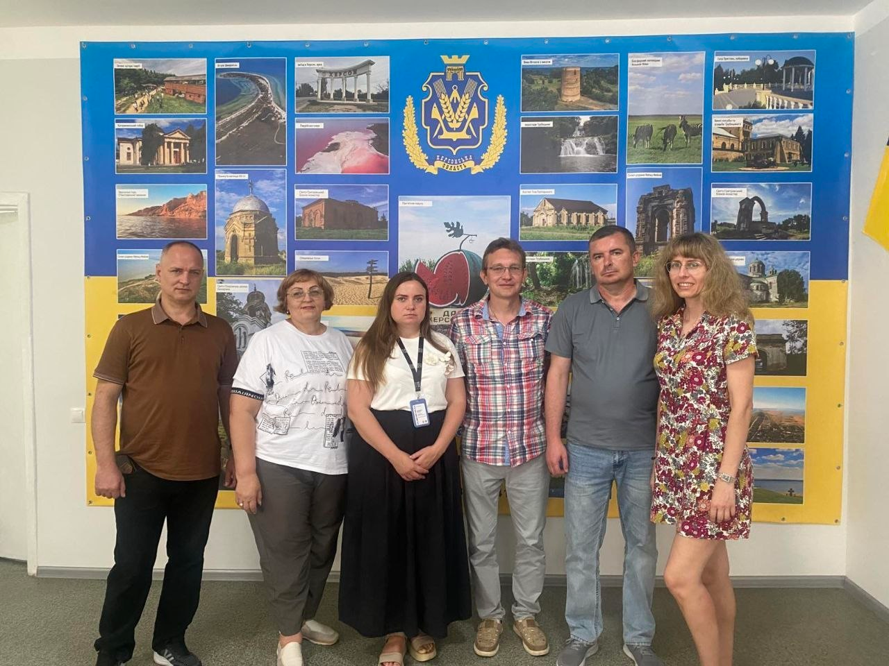 Регіональна координаторка громадської організації «Крим SOS» Світлана Корсік стоїть по центру (у білій блузці з бейджем та довгій чорній спідниці) разом із представниками Горностаївської та Рубанівської громад — усі учасники дебрифінгу щодо підтримки ВПО сфотографовані у ряд на тлі великого синьо-жовтого інформаційного стенда з гербом та краєвидами Херсонської області.