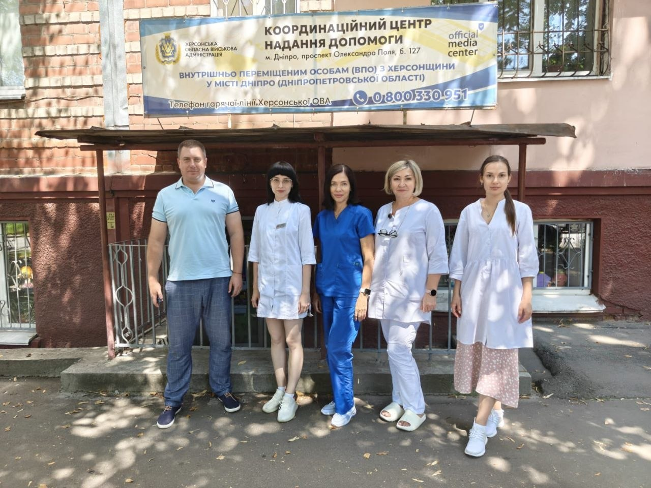 Команда медиків Луганської обласної клінічної лікарні та представник центру стоять біля входу до Координаційного центру допомоги ВПО з Херсонщини в Дніпрі. Чотири жінки-лікарки вбрані у білу та синю медичну уніформу, поруч — представник центру вбраний у блакитну футболку-поло, сірі штани та сині кросівки. Над ними висить інформаційний банер установи.