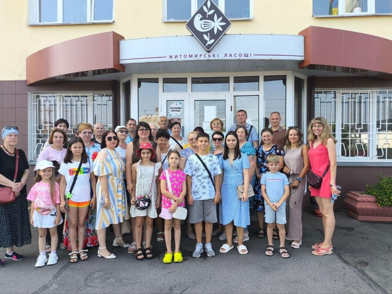 Групове фото переселенців з Херсонщини, організоване координаційним центром «Вільні разом», біля входу до кондитерської фабрики «Житомирські ласощі» у Житомирі. Велика група дорослих та дітей позирує для спільного знімка на тлі скляних дверей та фірмової вивіски підприємства після завершення екскурсії виробництвом.