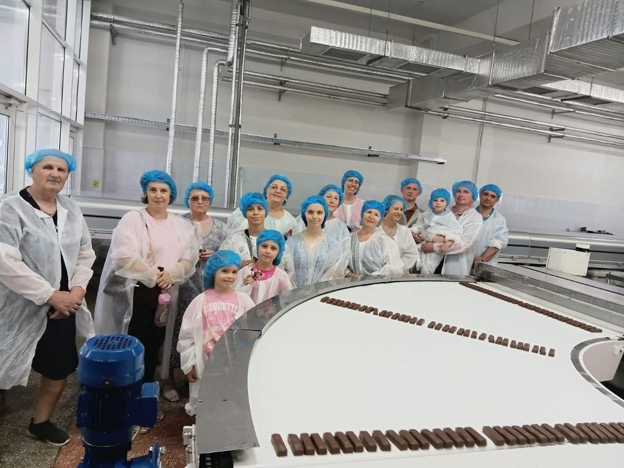 Переселенці з Херсонщини під час екскурсії у виробничому цеху кондитерської фабрики «Житомирські ласощі». Група дорослих та дітей у захисних білих халатах і блакитних шапочках стоїть біля конвеєрної стрічки, на якій рухаються ряди шоколадних цукерок або батончиків.