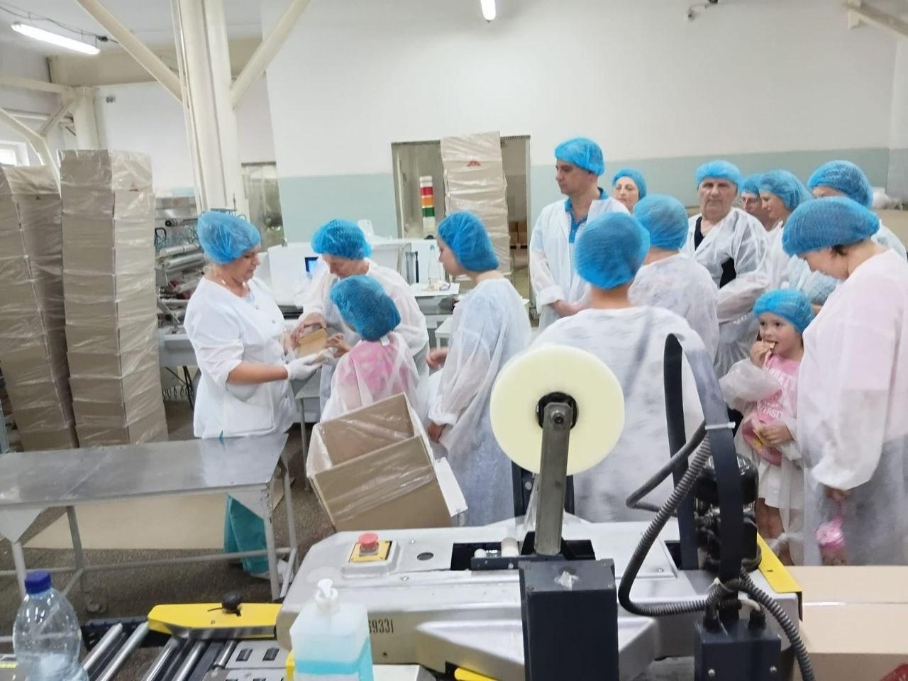 Переселенці з Херсонщини під час екскурсії пакувальним цехом кондитерської фабрики «Житомирські ласощі». Працівниця підприємства у білому халаті та рукавичках демонструє групі відвідувачів у захисному одязі процес пакування готової продукції в картонні коробки.