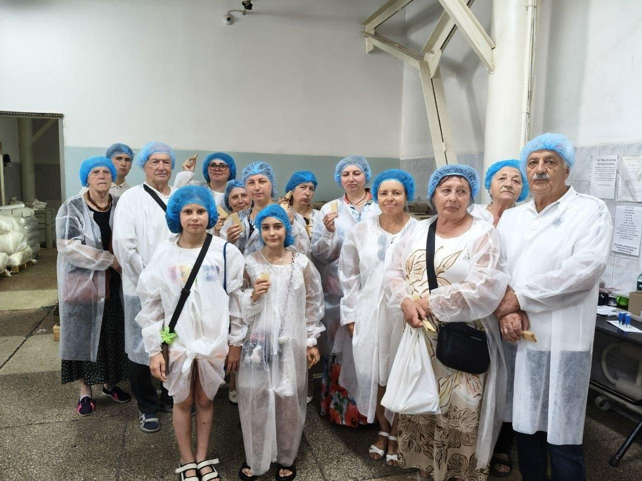 Група переселенців з Херсонщини в захисних одноразових халатах та шапочках під час дегустації солодощів у виробничому цеху кондитерської фабрики «Житомирські ласощі». Дорослі та діти стоять разом у приміщенні фабрики, тримаючи в руках свіжі кондитерські вироби.