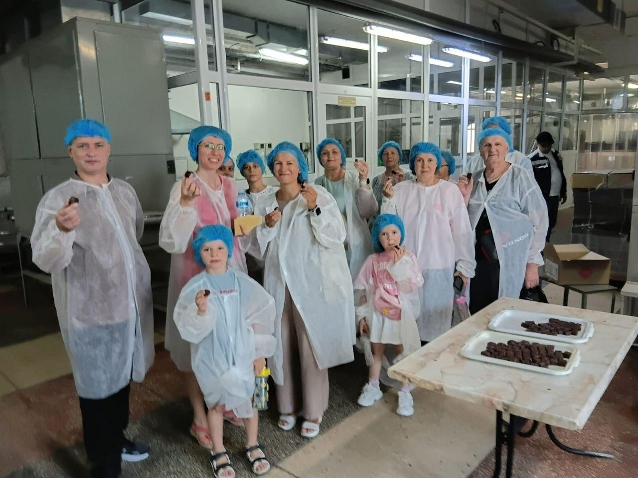 Група переселенців з Херсонщини під час дегустації шоколадних цукерок у кондитерському цеху фабрики «Житомирські ласощі». Дорослі та діти в захисних халатах і блакитних шапочках стоять біля столу з розкладеними солодощами та тримають цукерки в руках.