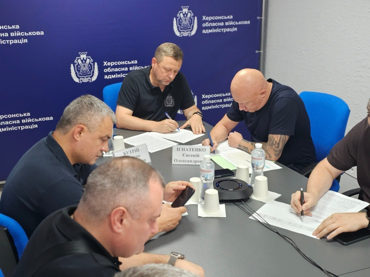 Учасники наради від Херсонщини під час онлайн-обговорення комплексних планів стійкості регіонів. За столом на тлі синього банера «Херсонська обласна військова адміністрація» працюють посадовці: у центрі — перший заступник начальника ХОВА Дмитро Бутрій, праворуч від нього — заступник начальника ХОВА Євгеній Ігнатенко, а праворуч Ігнатенка на передньому плані частково видно директора Департаменту з питань цивільного захисту та оборонної роботи Херсонської ОДА Євгена Пісоцького, який робить записи в документах.
