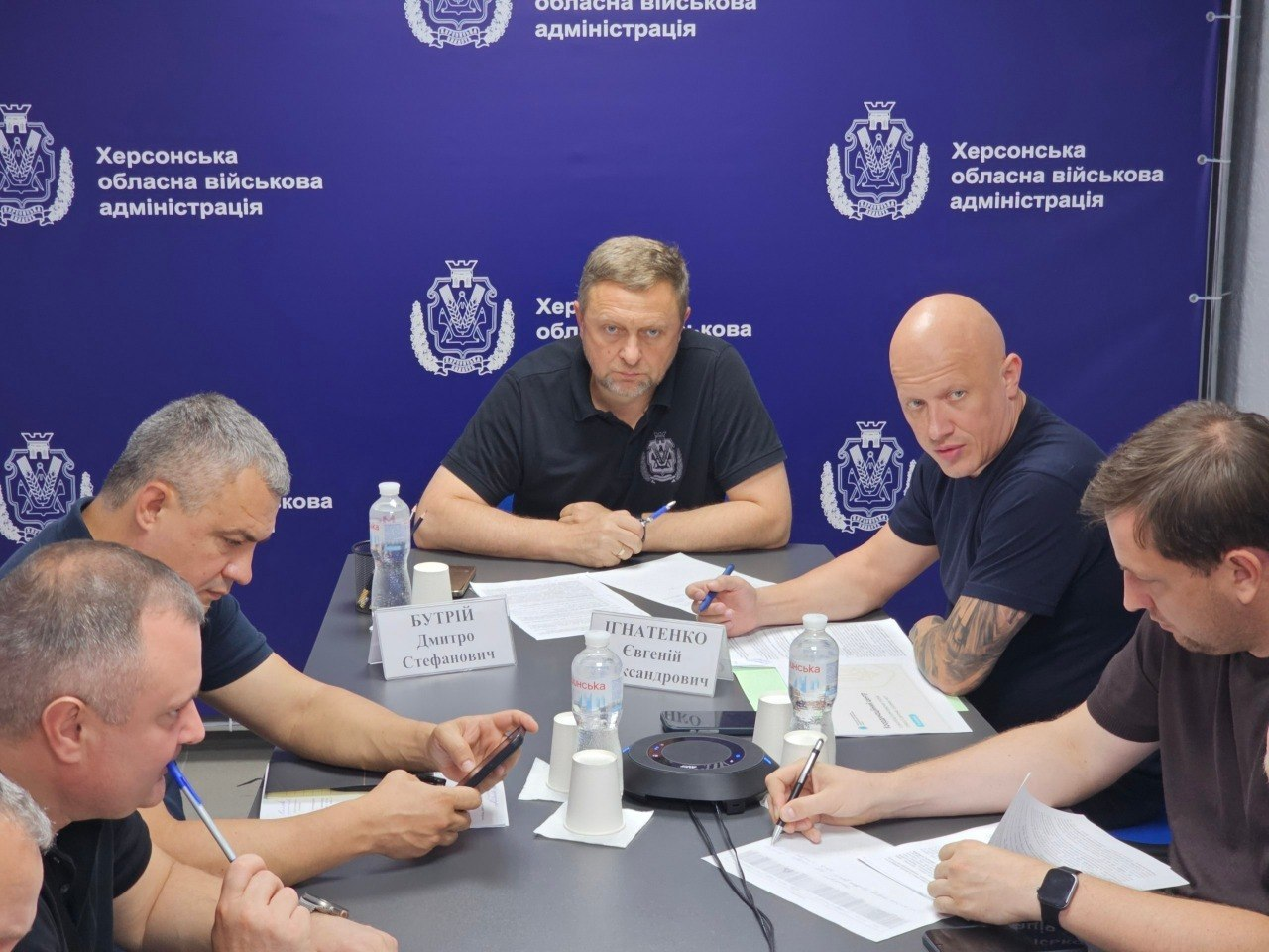 Учасники наради від Херсонщини під час онлайн-обговорення комплексних планів стійкості регіонів. За столом на тлі синього банера «Херсонська обласна військова адміністрація» сидять посадовці: у центрі — перший заступник начальника ХОВА Дмитро Бутрій, праворуч від нього — заступник начальника ХОВА Євгеній Ігнатенко, а праворуч Ігнатенка на передньому плані — директор Департаменту з питань цивільного захисту та оборонної роботи Херсонської ОДА Євген Пісоцький. Також за столом присутні інші колеги, які працюють із документами.