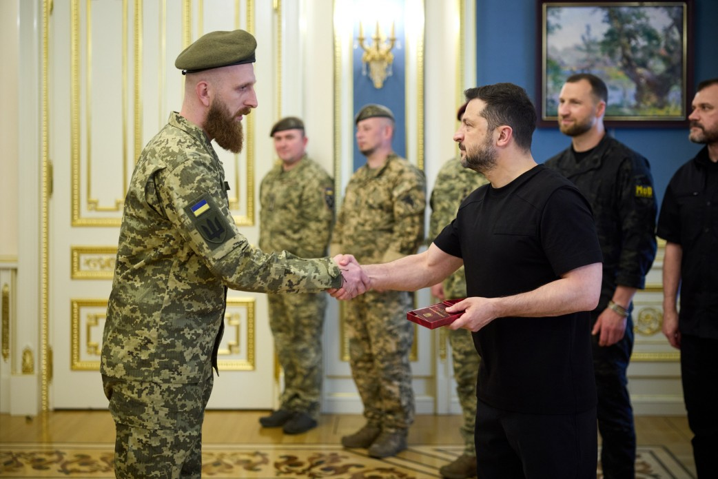 Президент Володимир Зеленський у день професійного свята — Дня Військ зв’язку та кібербезпеки ЗСУ — тисне руку українському військовослужбовцю під час вручення державних нагород. Офіцер із густою довгою каштановою бородою одягнений у камуфляжний однострій із нашивкою Державного Прапора та тризуба на правому рукаві, а на голові має маслиновий берет. Глава держави тримає в руці відкриту коробочку з відзнакою, а на задньому плані стоять інші військові та посадовці в офіційній залі.