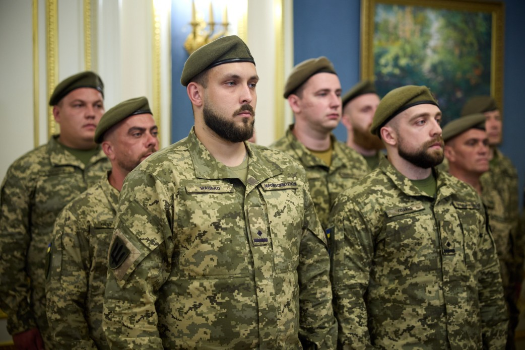 Група українських військовослужбовців Військ зв’язку та кібербезпеки ЗСУ в камуфляжній формі та зелених беретах стоїть у шерензі в залі Офісу Президента під час урочистостей до Дня Військ зв’язку та кібербезпеки. На передньому плані — офіцер із охайною темною бородою та нашивками на грудях. Праворуч від нього стоїть інший воїн із короткою бородою, а ліворуч та позаду — ще кілька побратимів, які зосереджено дивляться вперед. Позаду військових видно світлу стіну з колонами та картину в золотистій рамі.