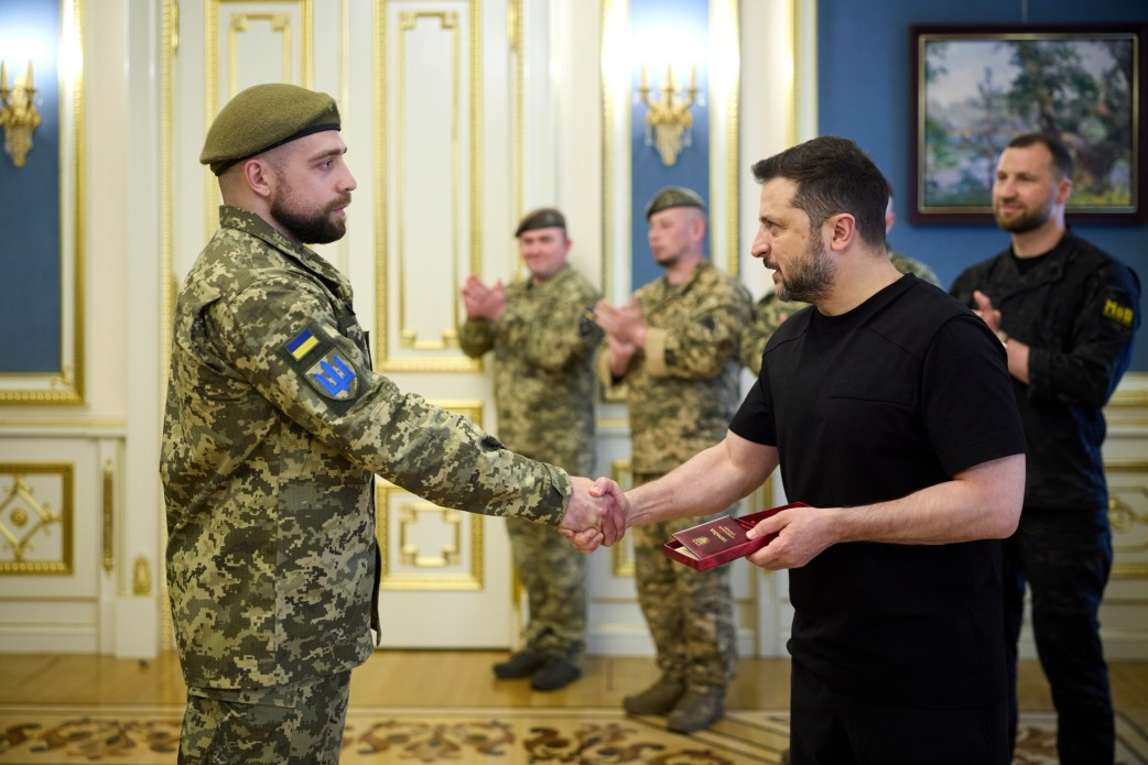 Президент Володимир Зеленський у день професійного свята — Дня Військ зв’язку та кібербезпеки ЗСУ — тисне руку українському військовослужбовцю під час вручення державних нагород. Офіцер із короткою темною бородою одягнений у камуфляжний однострій із нашивкою Державного Прапора та синьо-жовтого тризуба на правому рукаві, а на голові має маслиновий берет. Глава держави тримає в руці відкриту коробочку з відзнакою, а на задньому плані присутні плещуть у долоні в офіційній залі.