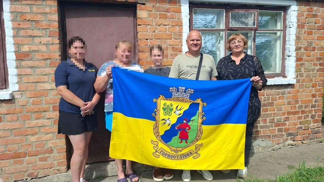 Начальник Горностаївської СВА Дмитро Ляхно та фахівці тримають прапор Горностаївки разом із родиною, повернутою з ТОТ, яка наразі проживає на Вінниччині. Вони стоять на тлі одноповерхового будинку з червоної цегли із зачиненими коричневими дверима та старим дерев'яним вікном із білою рамою; обличчя членів сім'ї розмиті з міркувань безпеки.