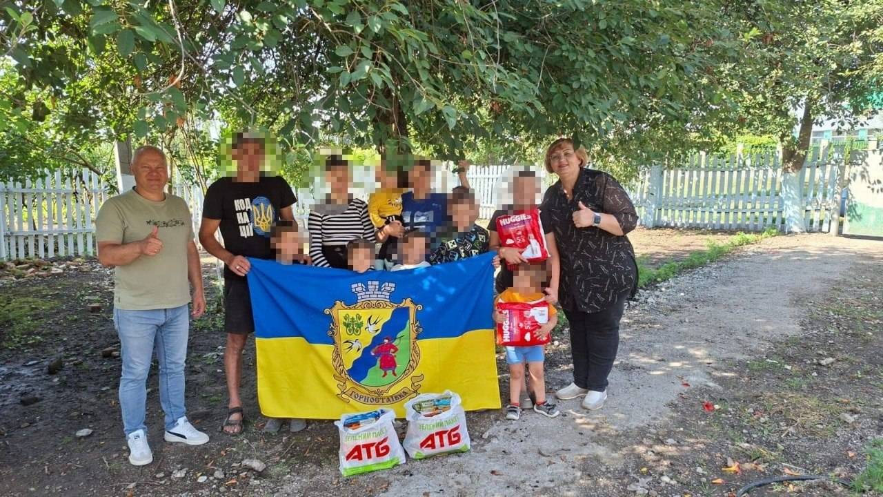 Начальник Горностаївської СВА Дмитро Ляхно та фахівці разом із багатодітною родиною, повернутою з ТОТ, яка наразі проживає на Вінниччині, тримають прапор Горностаївки на зеленому подвір'ї з деревами та білим парканом на задньому плані. На передньому плані на землі стоять пакунки з гуманітарною допомогою, а діти тримають у руках упаковки підгузків Huggies; обличчя членів родини розмиті з міркувань безпеки.
