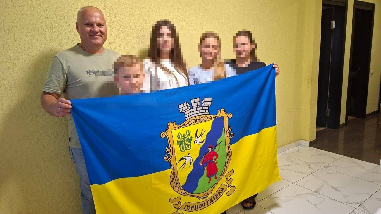 Начальник Горностаївської СВА Дмитро Ляхно та фахівці тримають прапор Горностаївки разом із родиною, повернутою з ТОТ, яка наразі проживає на Вінниччині. Вони стоять у приміщенні зі світло-жовтими стінами та білою плиткою на підлозі, а обличчя членів сім'ї розмиті з міркувань безпеки.