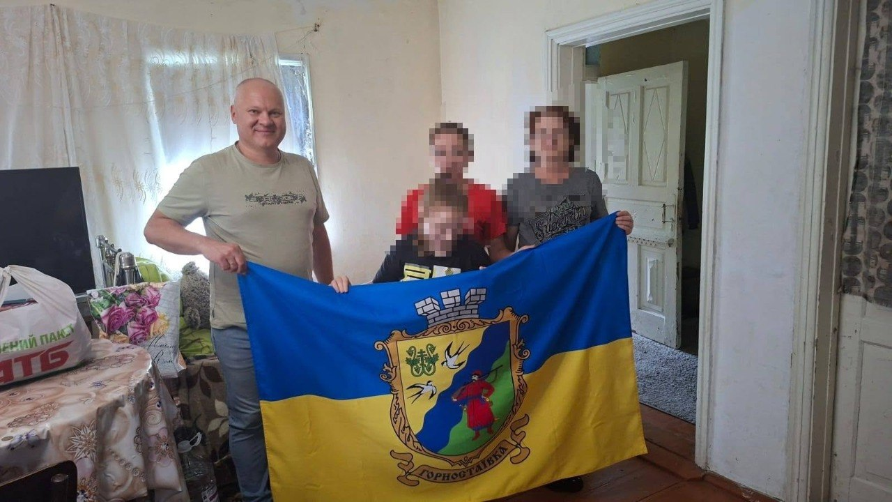 Начальник Горностаївської СВА Дмитро Ляхно та фахівці тримають прапор Горностаївки разом із родиною, повернутою з ТОТ, яка наразі проживає на Вінниччині. Вони стоять у кімнаті житлового будинку зі світлими стінами та відчиненими старими білими дверима на задньому плані. Ліворуч на застеленому скатертиною столі стоять пакети та коробки з гуманітарною допомогою, а обличчя членів сім'ї розмиті з міркувань безпеки.
