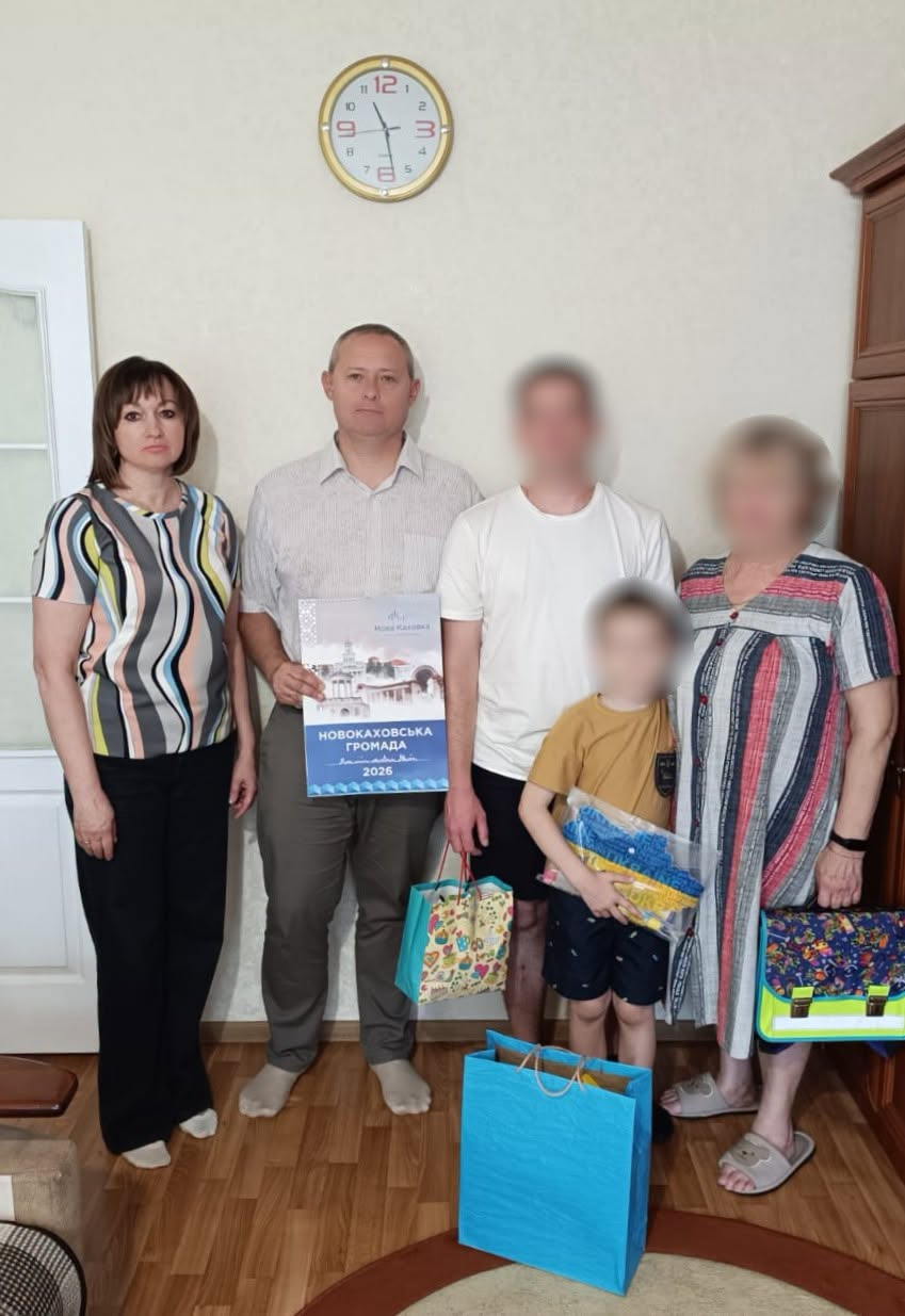 Начальник Каховської РВА Наталя Стаднік та начальник Новокаховської МВА Олег Тарабака, який тримає календар «Новокаховська громада 2026», відвідали родину, повернуту з ТОТ, яка знайшла прихисток у Кіровоградській області. Вони стоять у кімнаті з ламінатом на підлозі, світлими стінами, великим круглим годинником угорі та дерев'яною шафою праворуч; на передньому плані стоять блакитний і кольоровий подарункові пакети, а обличчя родини розмиті з міркувань безпеки.