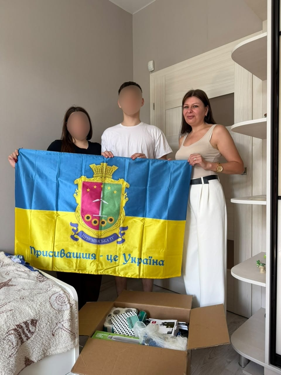 Начальниця служби у справах дітей Присиваської сільської ради Каховського району відвідала родину, яка знайшла прихисток в Одеській області після повернення з тимчасово окупованих територій. Вони разом стоять у світлій кімнаті біля полиць та тримають прапор громади із гербом і написом «Присивашшя — це Україна». На передньому плані стоїть відкрита картонна коробка з гуманітарною допомогою, де видно блокноти, термоси та інші речі, а обличчя родини розмиті з міркувань безпеки.
