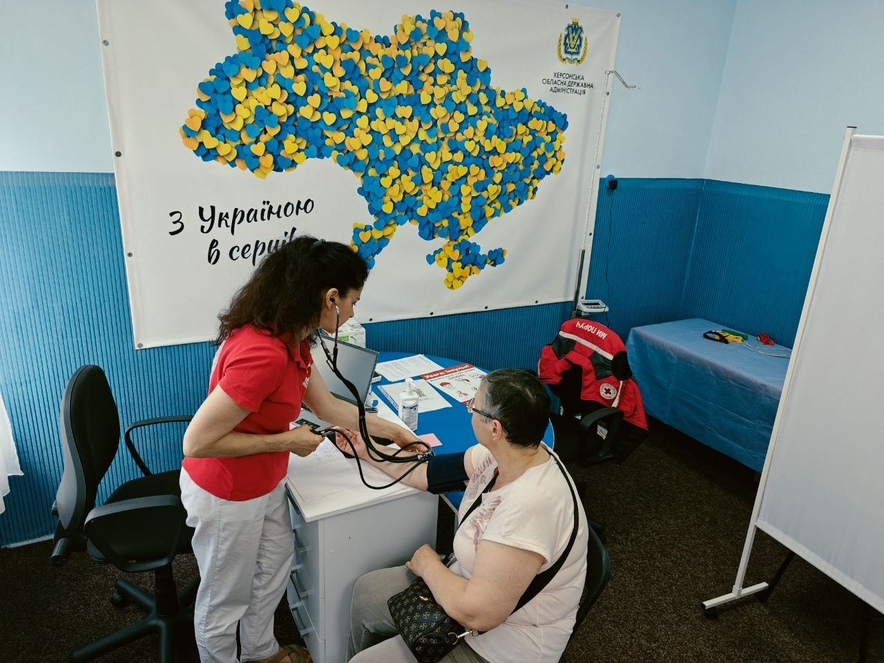 Лікар-терапевт КНП «Центр первинної медико-санітарної допомоги №3 м. Маріуполя» проводить медичний прийом, консультує жінку-переселенку та вимірює їй артеріальний тиск за допомогою тонометра в межах програми «Скринінг здоров’я 40+» у кабінеті гуманітарного центру «Вільні разом» у Хмельницькому. Вони сидять біля білого робочого столу, а на стільці поруч висить червоно-чорна куртка Товариства Червоного Хреста України. На задньому плані кабінету видно сині стіни, медичну кушетку та великий банер Херсонської ОДА із картою України з жовто-блакитних сердечок та написом «З Україною в серці!».