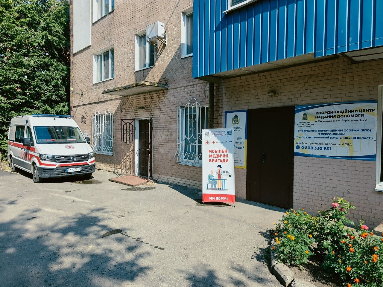 Вхід до Координаційного центру надання допомоги ВПО з Херсонщини у місті Хмельницький. Біля дверей будівлі встановлено інформаційні стенди, зокрема банер Товариства Червоного Хреста України «Мобільні медичні бригади», а ліворуч на подвір'ї припарковано автомобіль швидкої допомоги.