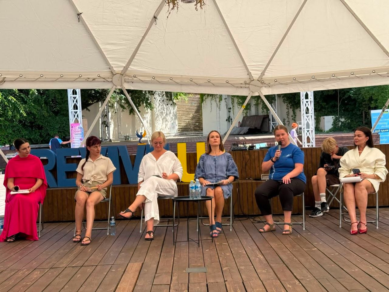 Спікерки фестивалю ODESA LIFE FEST беруть участь у панельній дискусії про розвиток та психологічну стійкість під білим купольним тентом в Одесі, до якої долучилися і ВПО з Херсонщини, і фахівці Верхньорогачицької громади Каховського району. На дерев'яній сцені на стільцях сидять шестеро жінок; одна з них у синій футболці говорить у мікрофон, інші тримають блокноти та слухають, а на тлі встановлено великі літери.