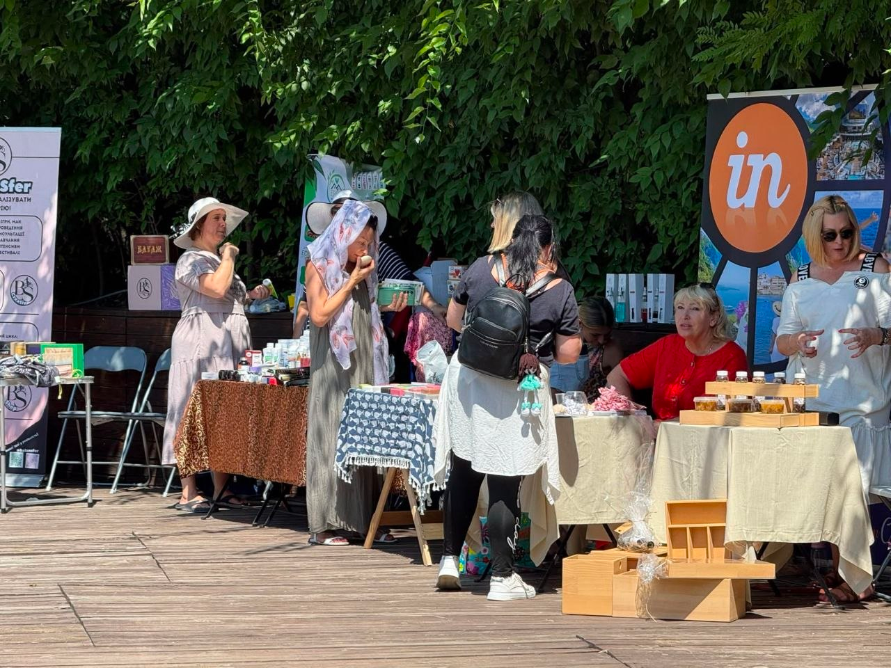 Учасники та відвідувачі фестивалю ODESA LIFE FEST, серед яких є ВПО з Херсонщини та фахівці Верхньорогачицької громади Каховського району, спілкуються біля виставкових ярмаркових столів просто неба. На столах розкладено різноманітну продукцію, косметику, інформаційні матеріали та солодощі, а поруч встановлено рекламні банери партнерських організацій. Жінки розглядають товари та беруть участь у нетворкінгу на тлі густої зеленої рослинності.