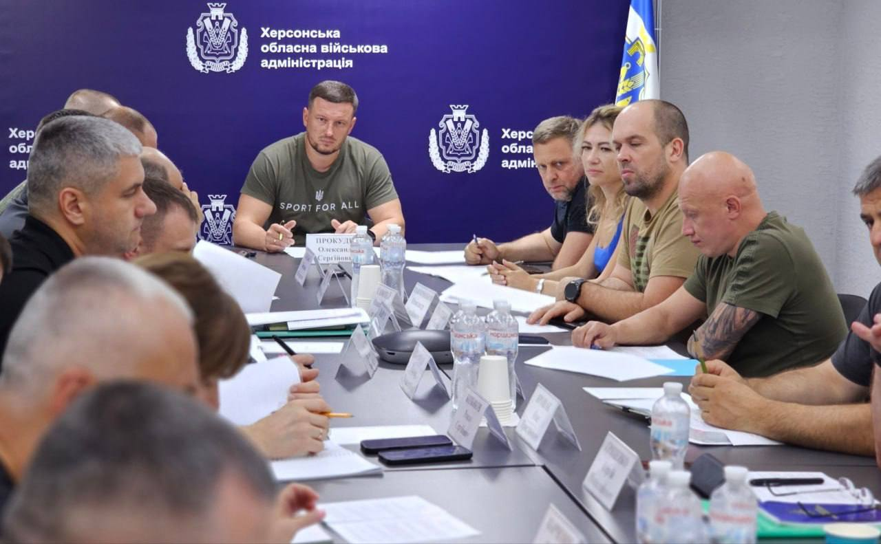 Олександр Прокудін проводить засідання Ради оборони Херсонщини, присвячене безпековій ситуації в Борозенській громаді, ухваленню рішення про розширення зони обов'язкової евакуації на села Красносільське та Чарівне., протидії дроновому терору РФ, посиленню бригад, результатам мобілізаційної кампанії та роботі ВЛК. Начальник ОВА сидить у центрі столу на тлі синього брендованого банера. Праворуч від нього за столом послідовно розташовані його заступники: перший заступник Дмитро Бутрій, заступниця Ольга Малярчук, заступники Олександр Толоконніков та Євгеній Ігнатенко. Навколо столу також зібралися інші посадовці ОДА, зокрема Департаменту здоров'я, представники військових відомств, Нацполіції та ТЦК та СП.