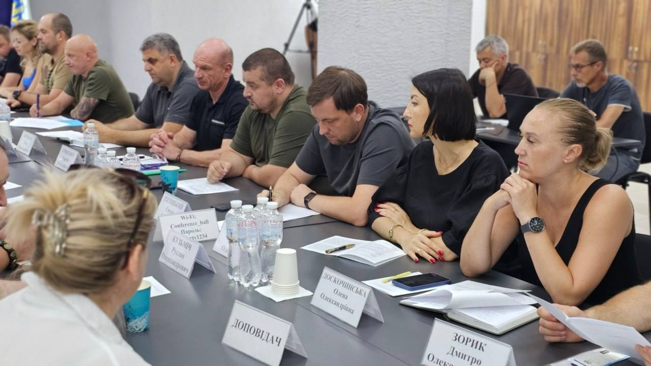 Посадовці ОДА та представники профільних служб беруть участь у засіданні Ради оборони Херсонщини. Учасники наради сидять уздовж довгого конференц-столу, вивчають папери, роблять нотатки та заслуховують доповіді щодо безпекової ситуації в області, результатів мобілізаційної кампанії, протидії дроновому терору РФ, посилення бригад та розширення зони обов'язкової евакуації населення.